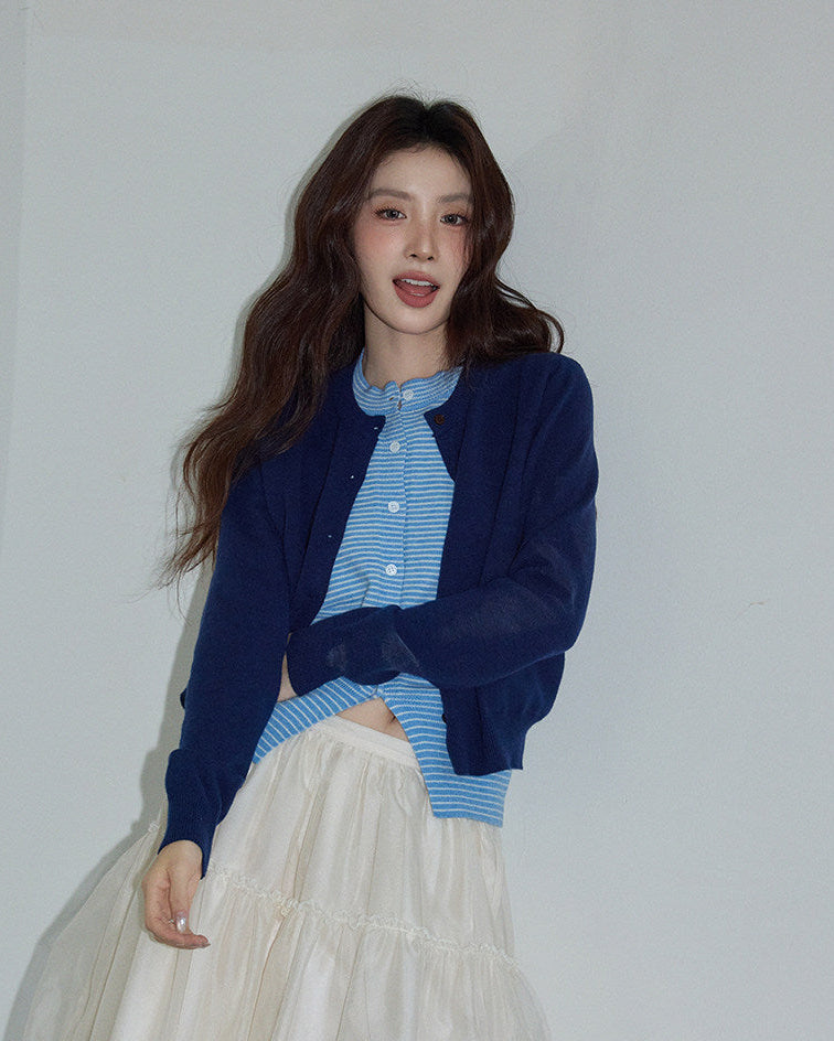 Woman wearing a blue cardigan over a striped shirt with a white skirt against a plain background