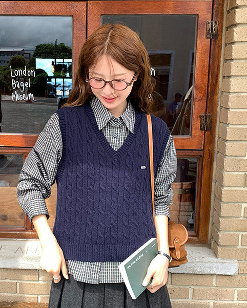 Woman standing outside a building holding a book, wearing a navy sweater vest and gray skirt.
