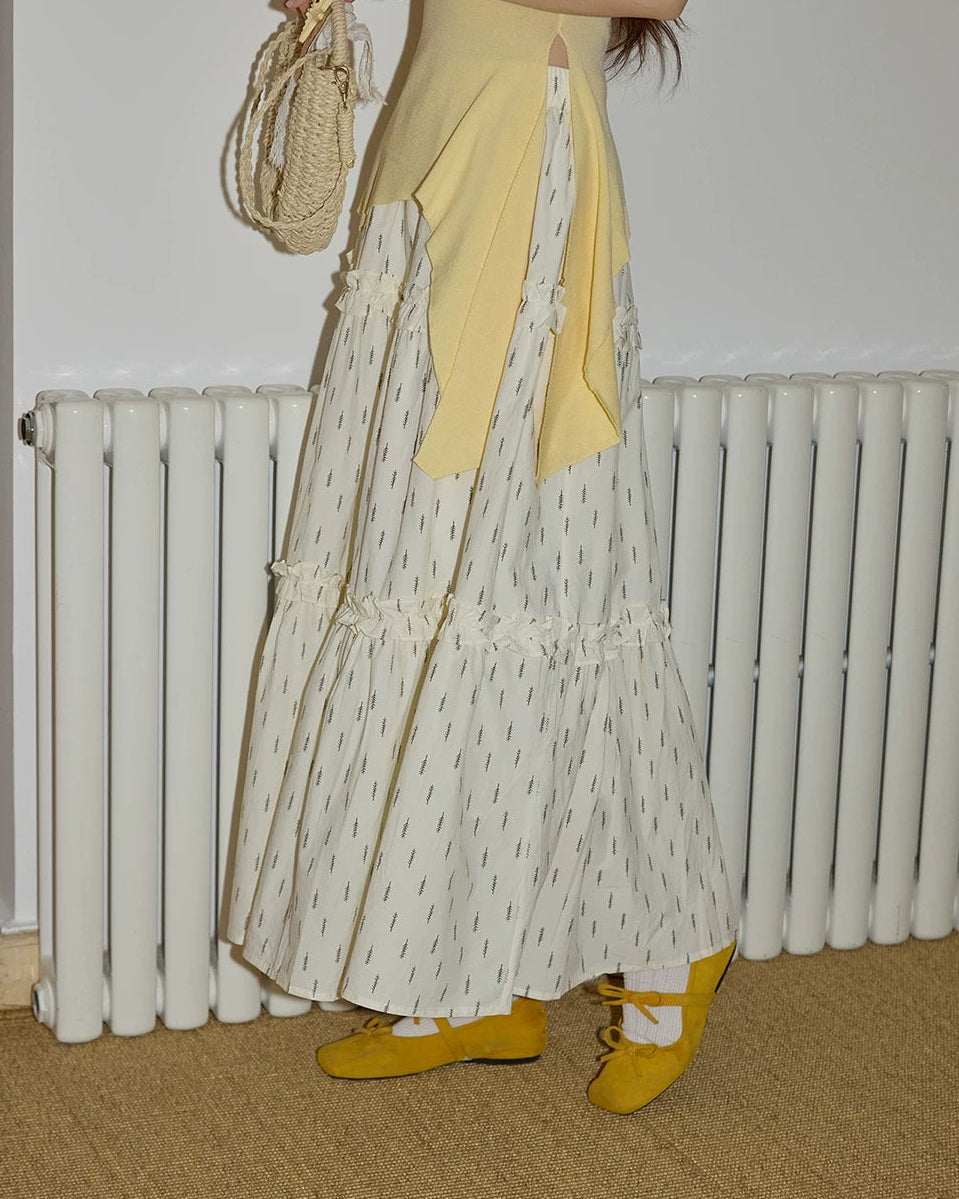 Woman wearing a yellow top and patterned skirt holding a straw bag indoors.