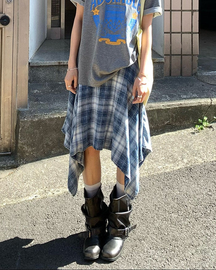 Person wearing a gray t-shirt with blue graphics and plaid skirt standing outside a building.