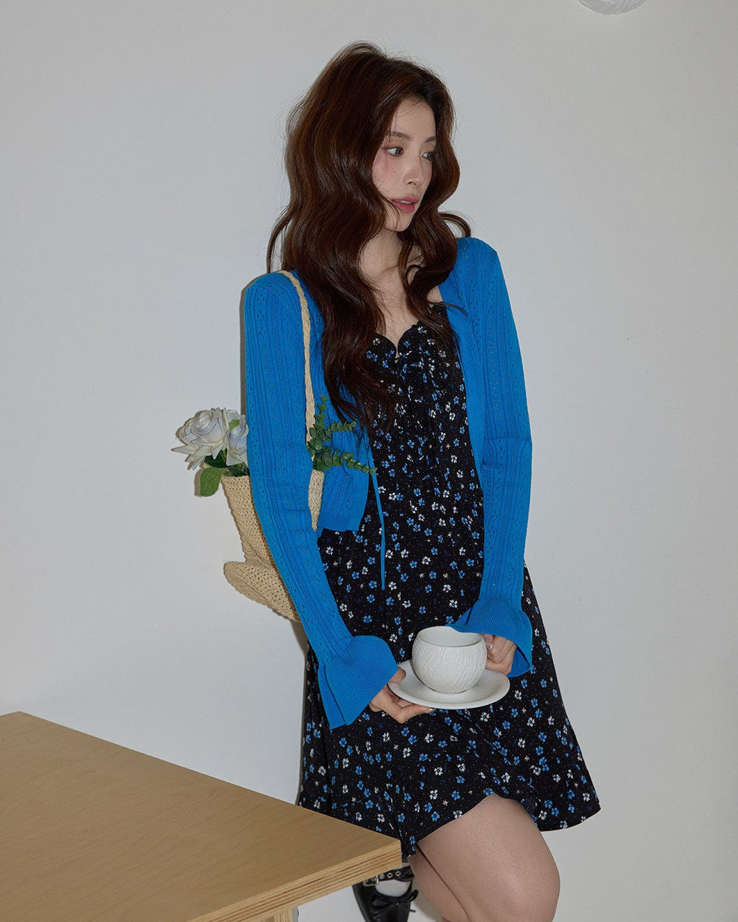 Woman in a blue cardigan holding a cup and saucer in a room with a table and flowers.